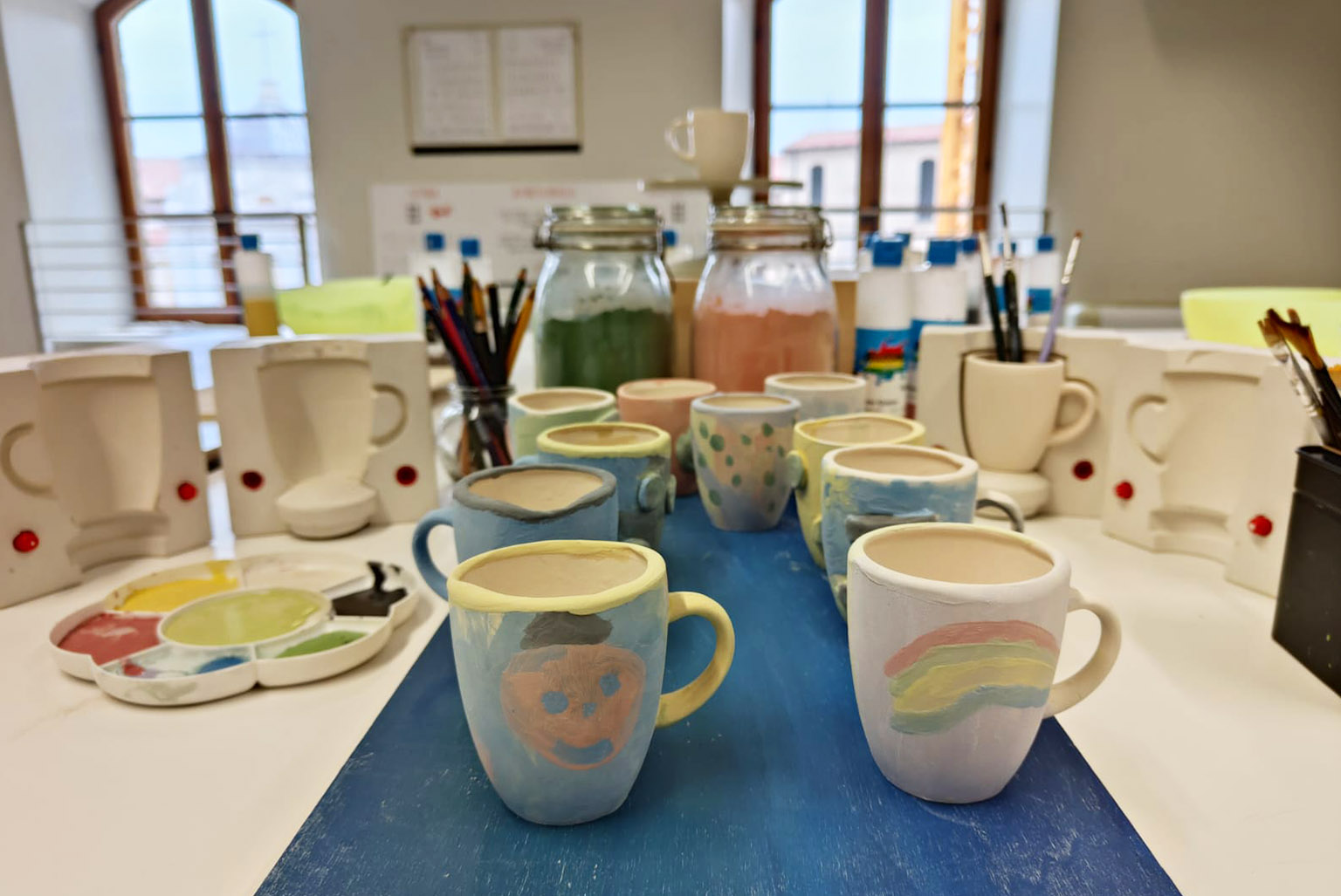Tavolo del laboratorio di ceramica con smalti, stampi, pennelli e alcune tazze colorate in fase di lavorazione.
