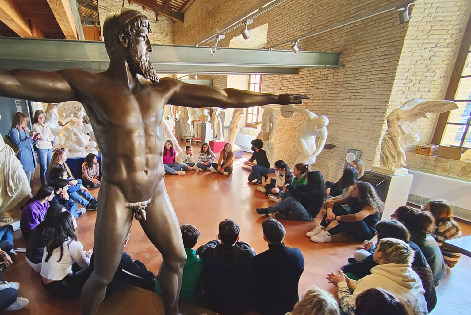 A group of young people sitting in a circle on the floor in the first room of the Museum. In the foreground, on the left, is the statue of Poseidon.