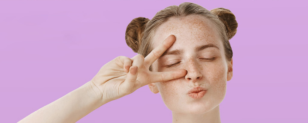 Ragazza con capelli raccolti in due chignon che fa il segno della vittoria con le dita davanti agli occhi e manda un bacio, su sfondo viola pastello.