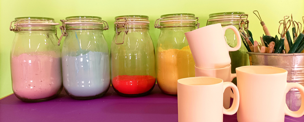 Su un piano viola tre barattoli di smalti colorati, contenitore con mirette e in primo piano tazzine di ceramica ancora da smaltare.