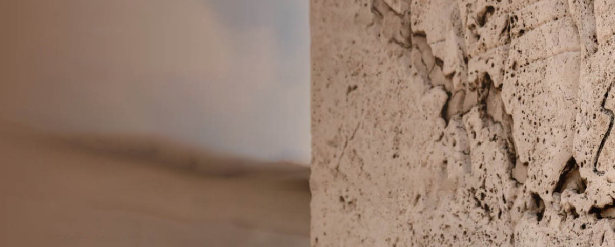 Superficie di pietra lavorata con crepe e segni incisi in primo piano; sullo sfondo sfocato, un paesaggio morbido e atmosferico che sfuma tra terra e cielo, creando un contrasto tra materia ruvida e orizzonte delicato.